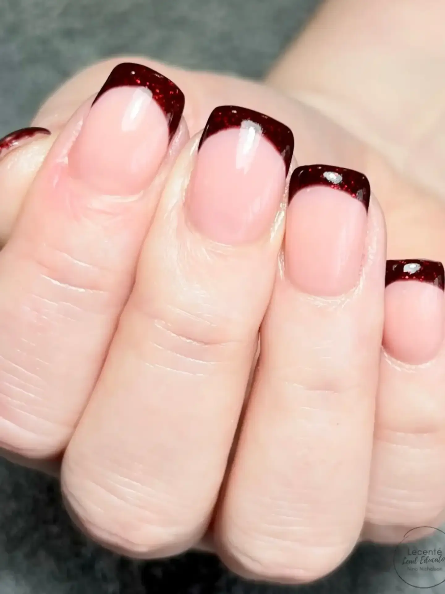 A close-up of a hand with neatly manicured nails featuring a nude base and dark burgundy glitter French tips. The nails are square-shaped and have a glossy finish.