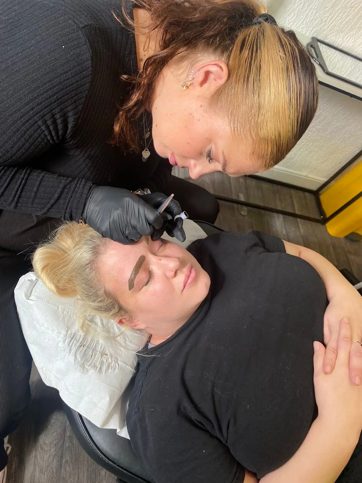 A beauty technician in black gloves carefully tattoos the eyebrows of a blonde woman lying on a treatment couch with her eyes closed. Both women wear black shirts in a well-lit salon setting.