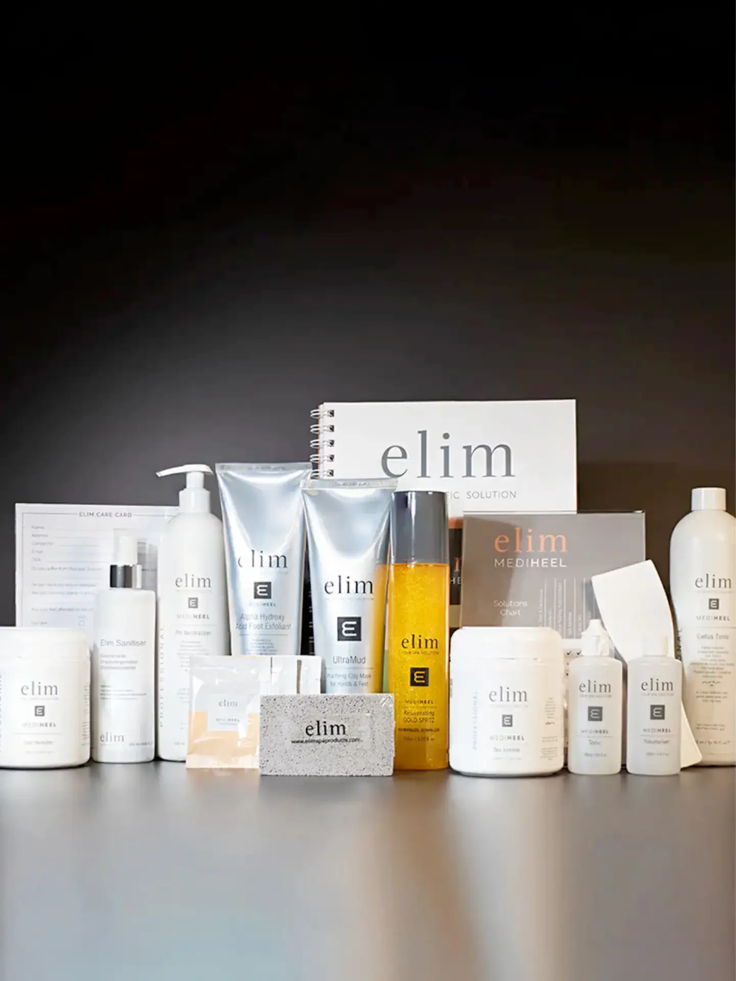 A display of various Elim-branded skincare products, including bottles, jars, tubes, a pumice stone, and a booklet, all arranged on a flat surface against a dark background.