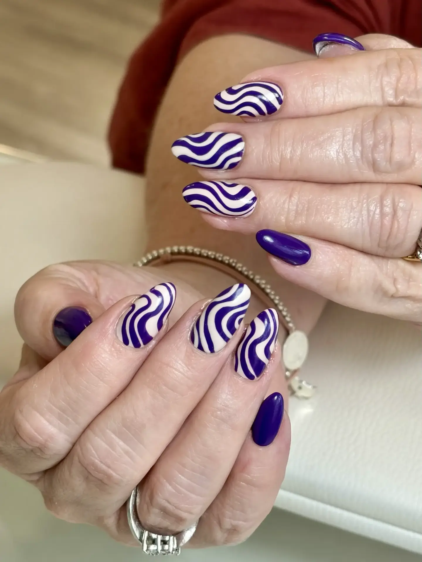 Close-up of hands with manicured nails, featuring a mix of solid purple nails and nails with bold purple and white wavy swirl designs. The hands rest on a light surface, and the person is wearing a silver bracelet.