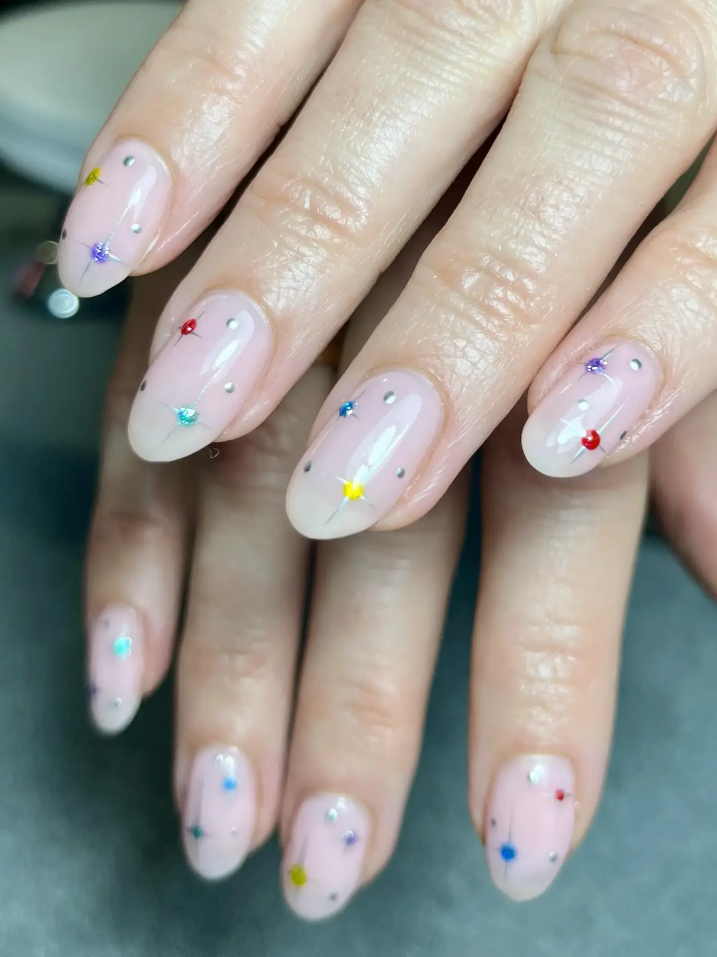 A close-up of hands with almond-shaped nails featuring a translucent nude base and small, colourful rhinestones and dots scattered across each nail for a delicate, festive look.