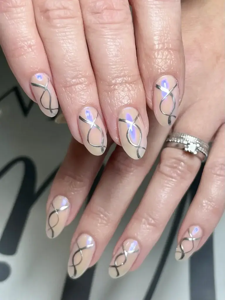 Close-up of hands with almond-shaped nails featuring a nude base, iridescent accents, and abstract black line designs. The person is wearing silver rings, including one with small stones and a flower detail.