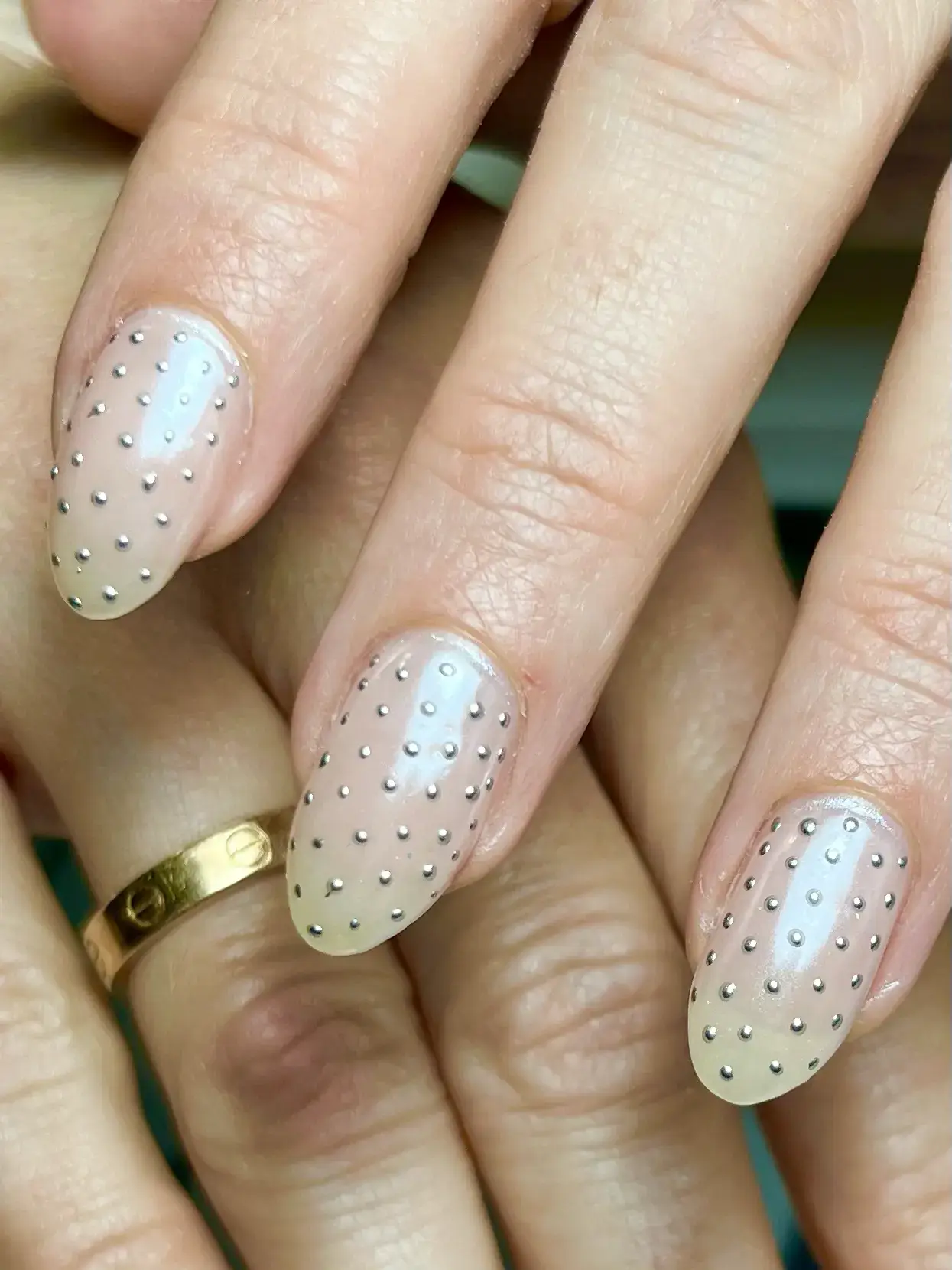A close-up of hands with almond-shaped nails painted in a nude shade, decorated with evenly spaced small silver studs. One hand features a gold ring on the ring finger.