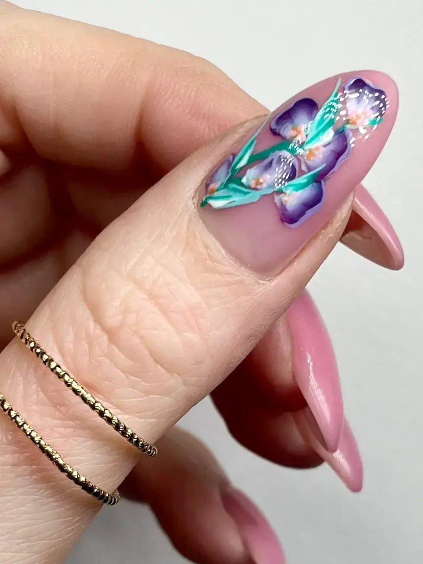 A close-up of a hand with long, almond-shaped nails; the thumb nail features a detailed purple floral design with green leaves, whilst the other nails are painted glossy pink. The person wears two thin gold rings on their finger.