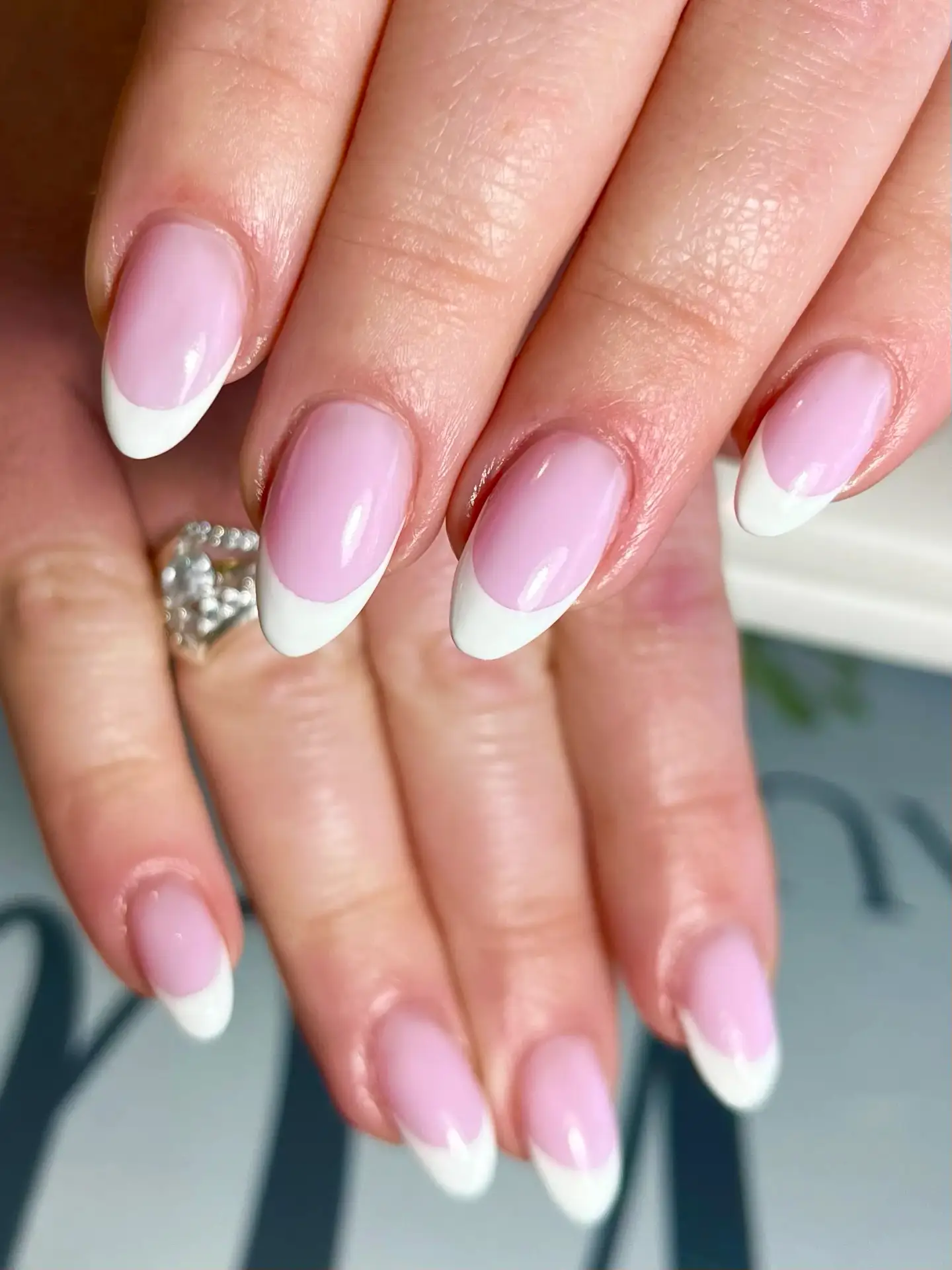Close-up of hands with almond-shaped nails featuring a classic French manicure with pale pink bases and white tips. A silver ring with clear stones is visible on one finger.