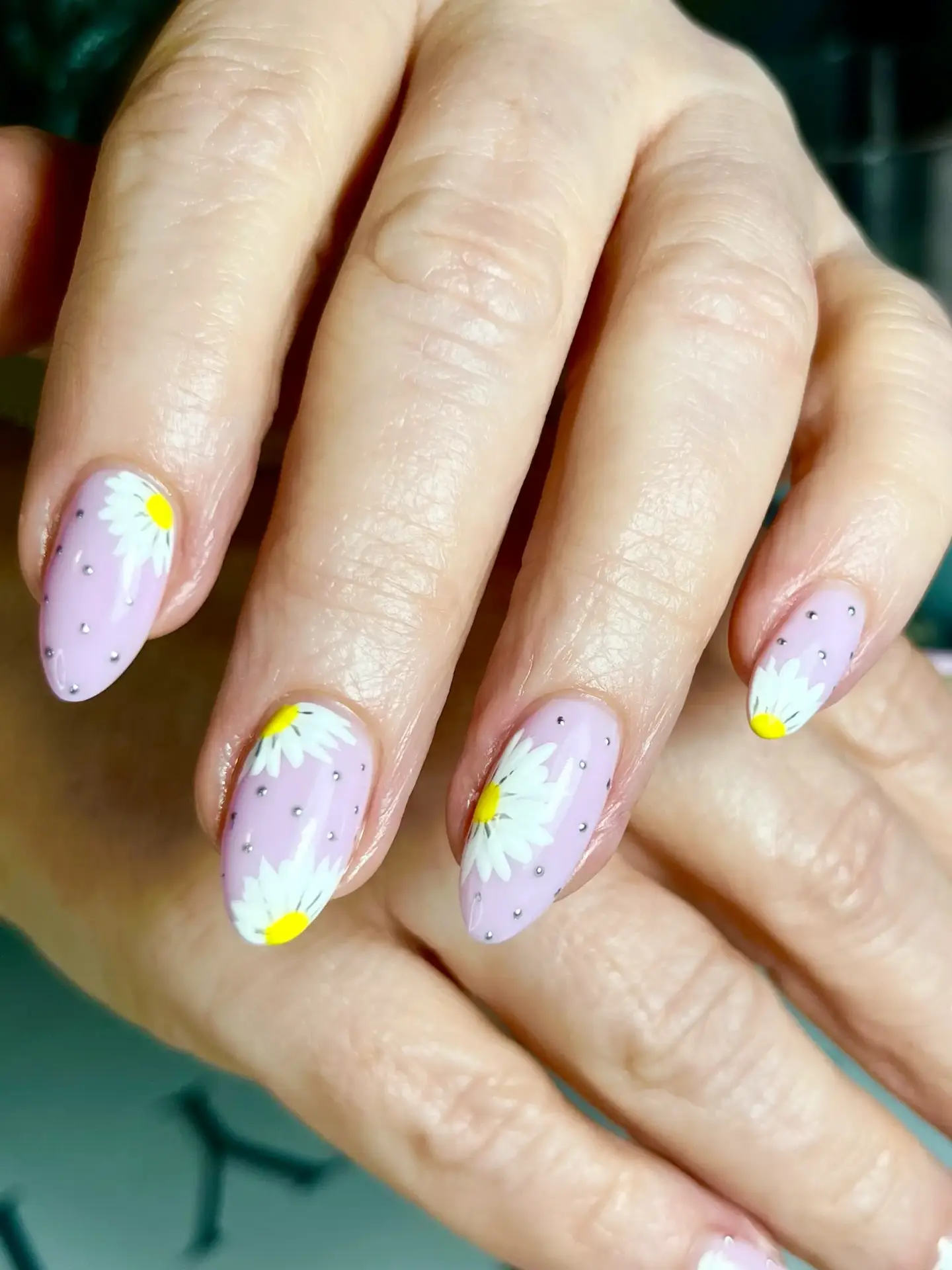 A close-up of hands with almond-shaped nails painted light pink, featuring white daisy flower designs with yellow centres and small silver dots as accents.