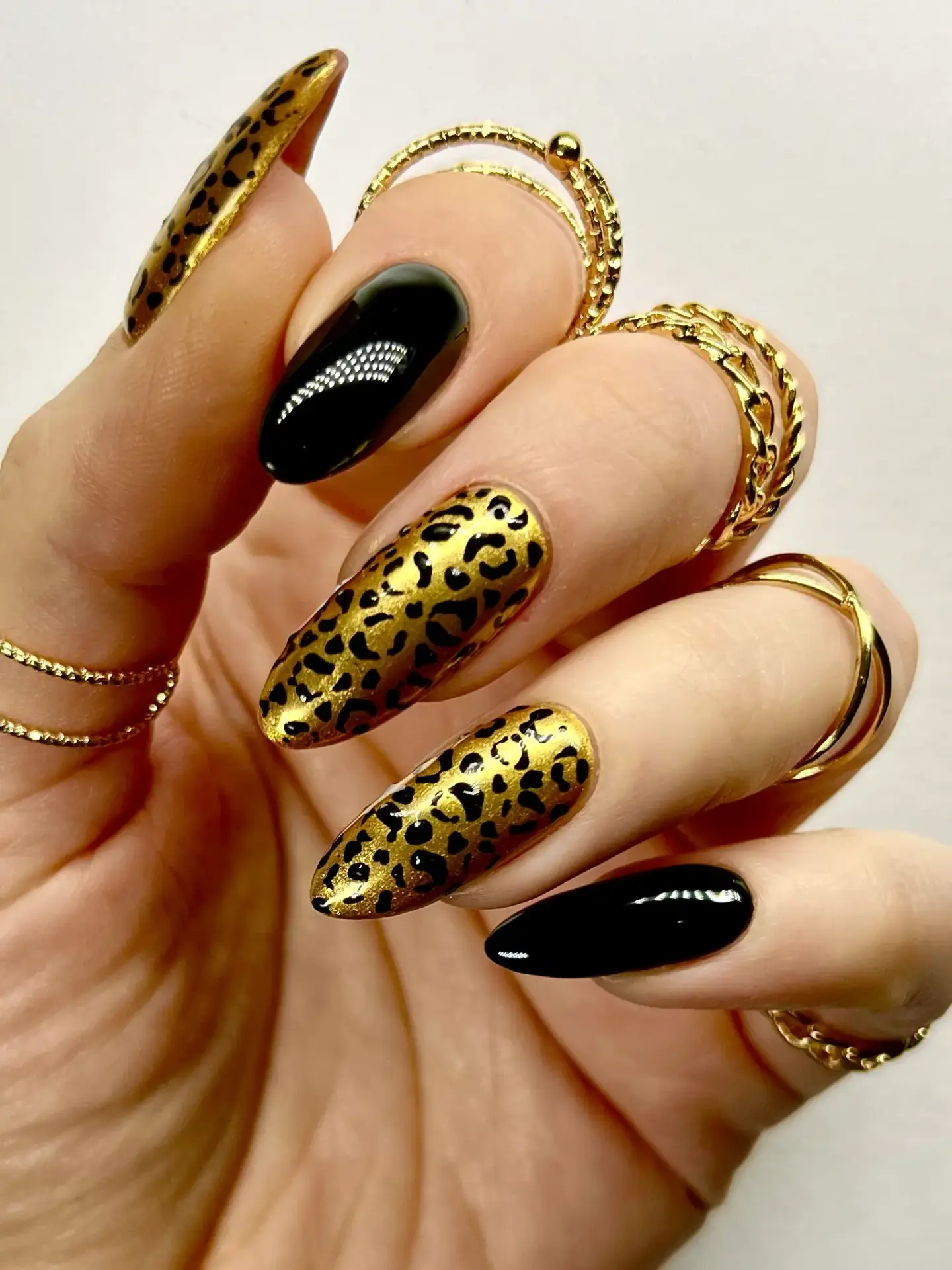 A hand with long, almond-shaped nails featuring black varnish and gold leopard print designs, adorned with multiple gold rings on each finger against a white background.