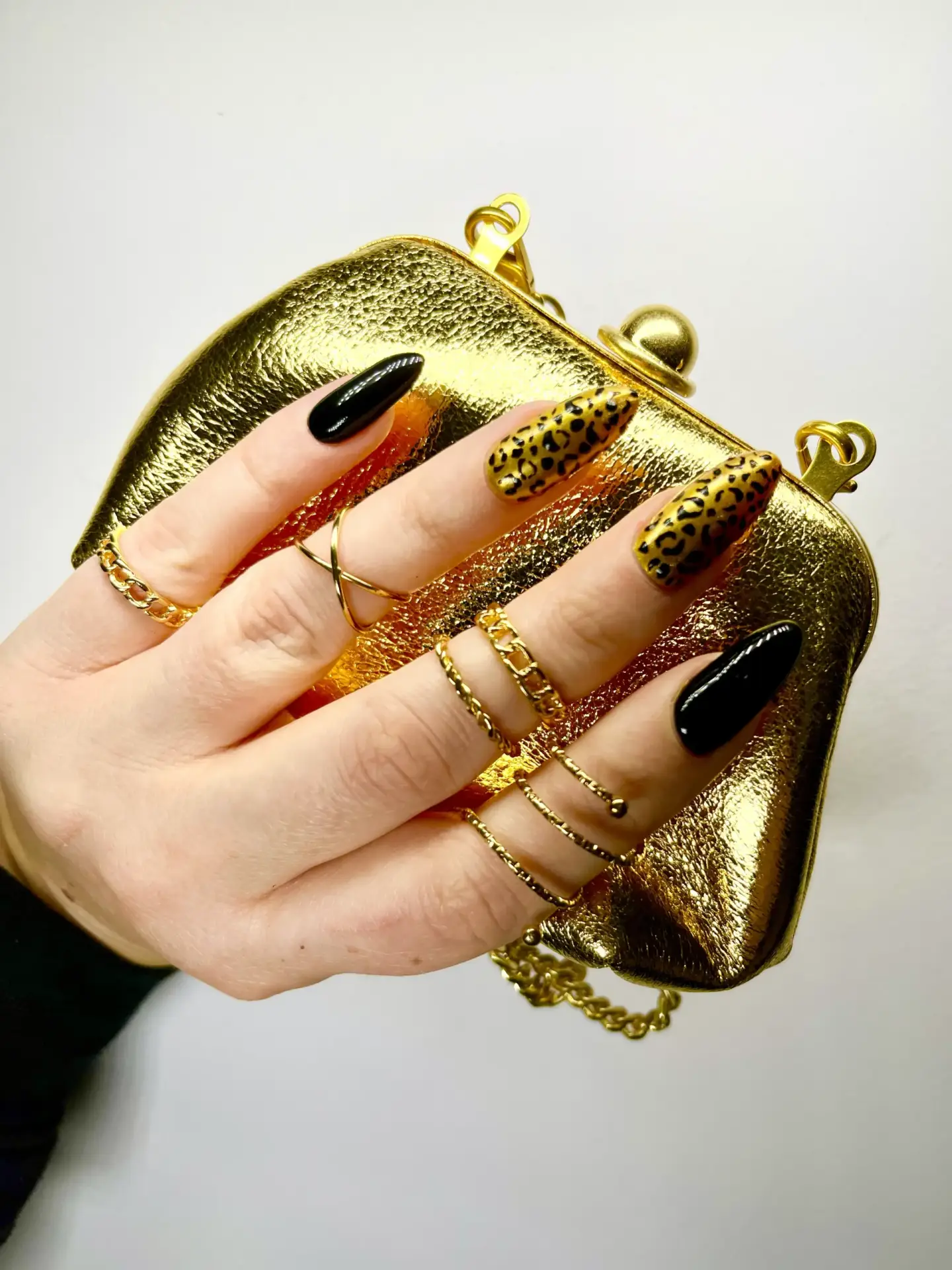 A hand with long, pointed, black and leopard-print nails holds a shiny gold handbag. The hand is adorned with several delicate gold rings and chain rings. The background is plain white.