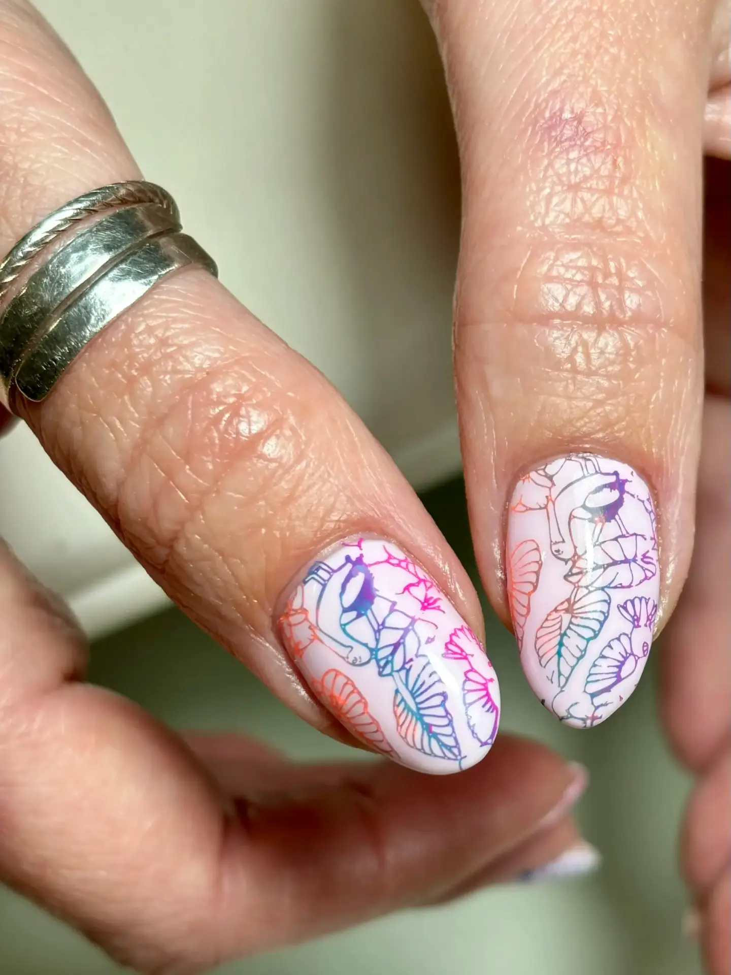 Close-up of two fingernails with pastel pink varnish, decorated with colourful abstract line art of faces and leaves. The person is wearing two silver rings on one finger.