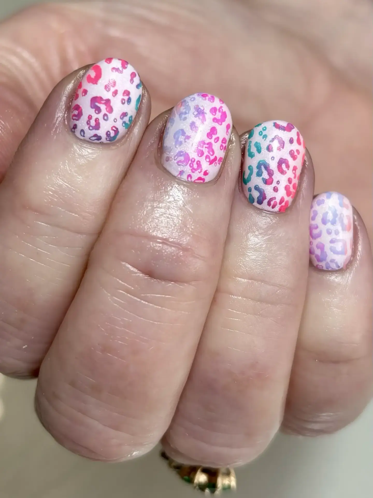 A close-up of a hand with short nails painted in a pastel ombré gradient, decorated with colourful leopard print patterns in pink, purple, blue, and red.