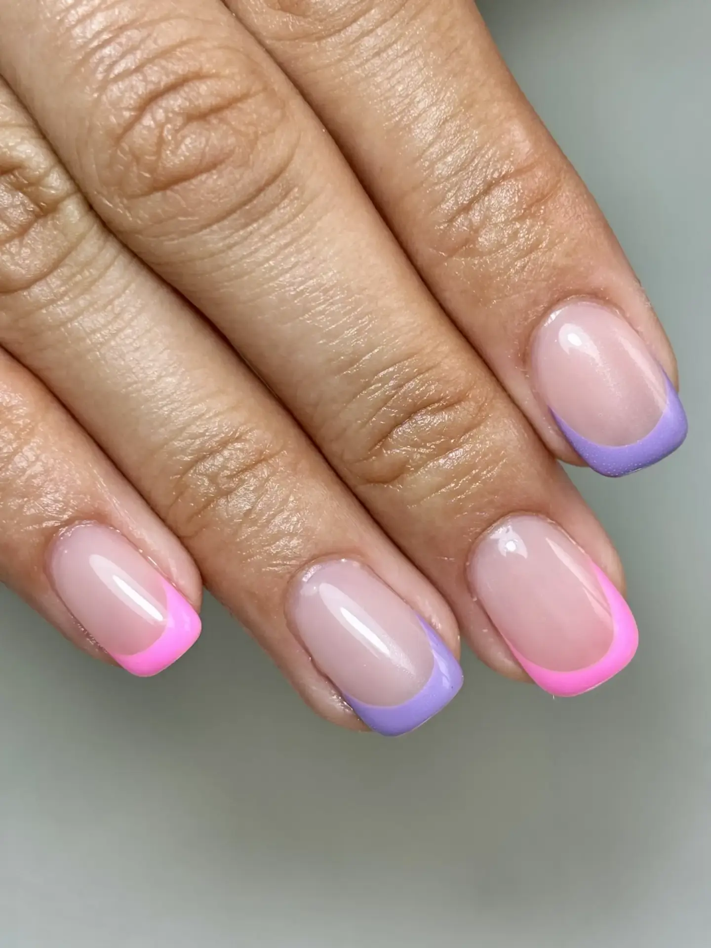 A hand with manicured nails featuring a French tip design: two nails have pastel purple tips and two have bright pink tips, whilst the rest of the nail is natural and glossy.