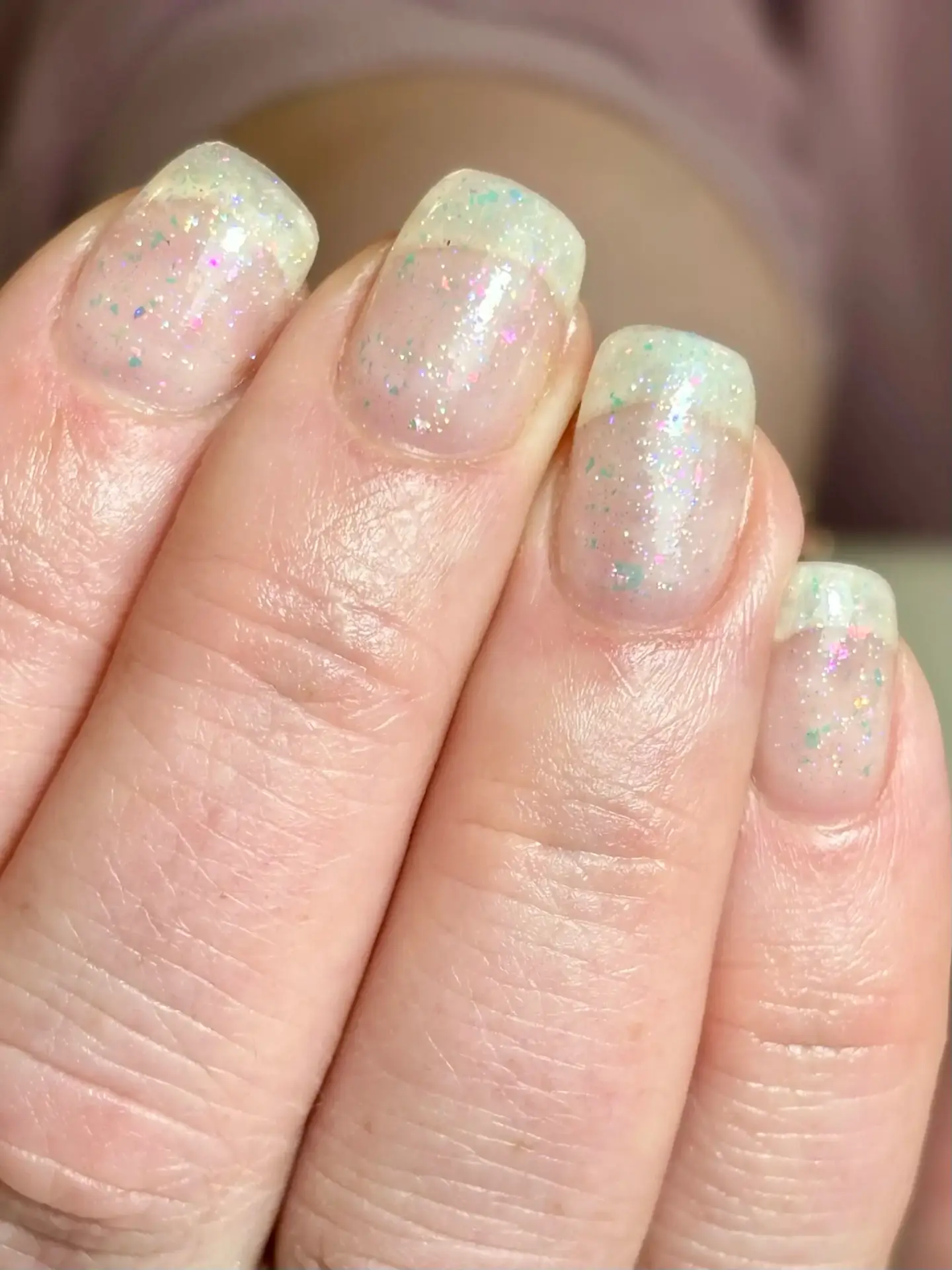A close-up of a hand with short, natural nails painted in a sheer, iridescent varnish with fine, multicoloured glitter. The skin tone is fair and the background is softly blurred.