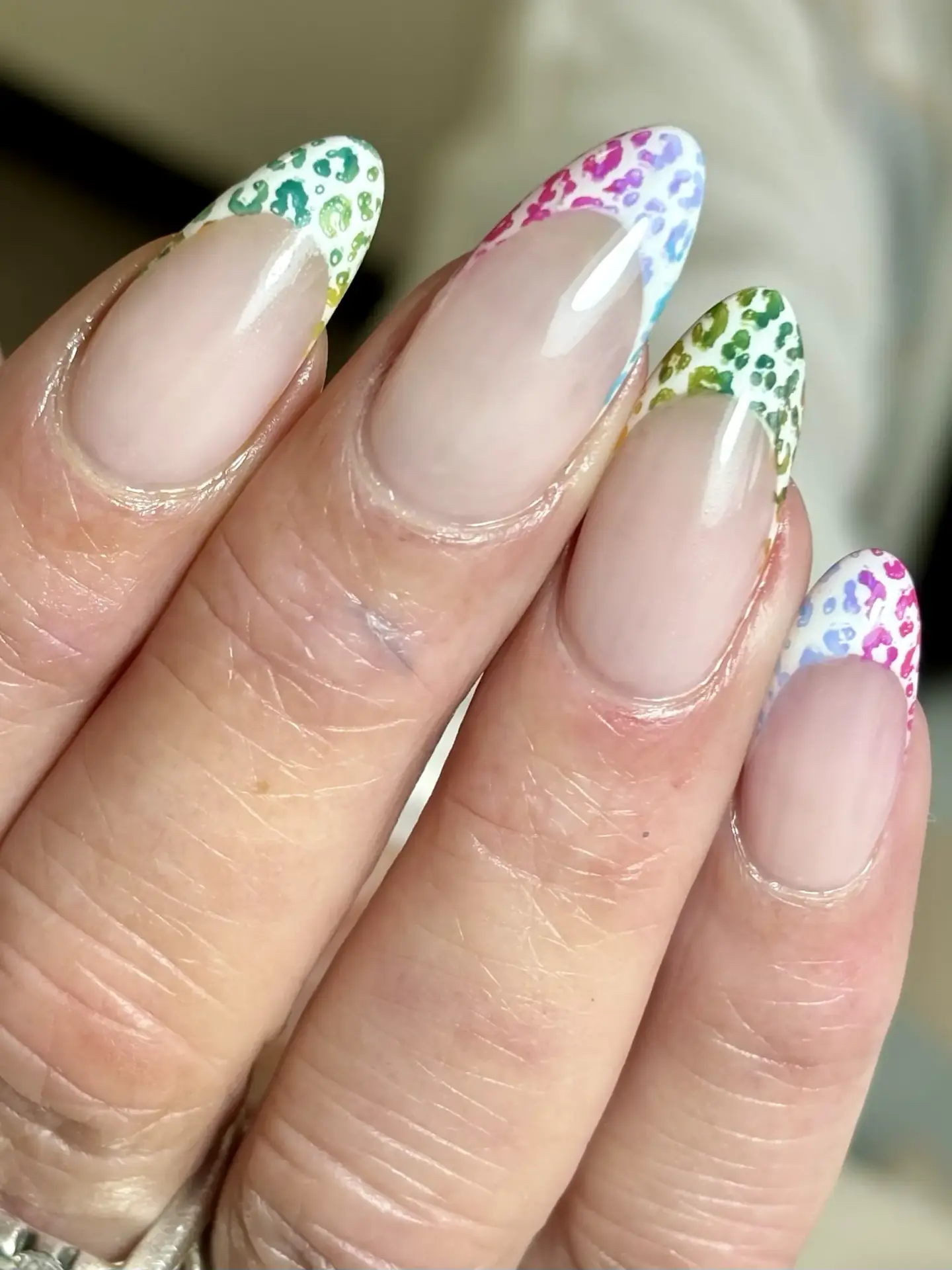 A hand with almond-shaped nails featuring a French tip design with multicoloured leopard print patterns on each tip, set against a neutral, glossy base.