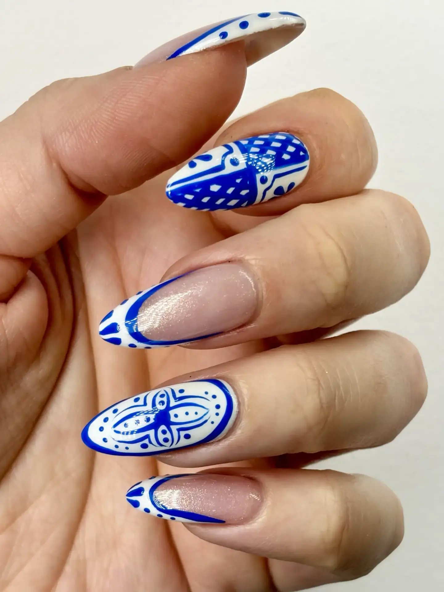A hand with long almond-shaped nails features intricate blue and white geometric and floral nail art designs on three nails, whilst two nails have a natural base with minimal blue accents.