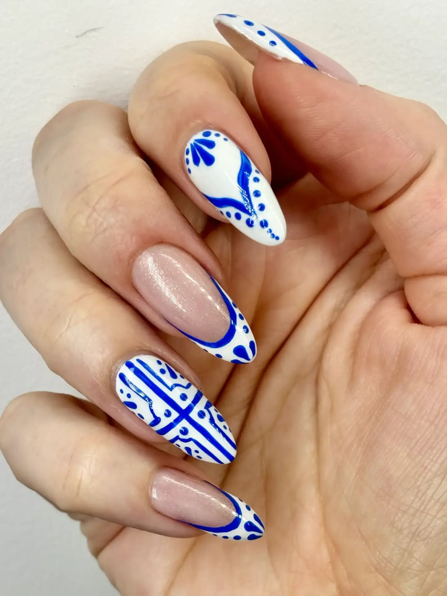 A hand with almond-shaped nails features a mix of nude polish and intricate blue and white patterns, including dots, lines, and geometric designs, on selected nails.