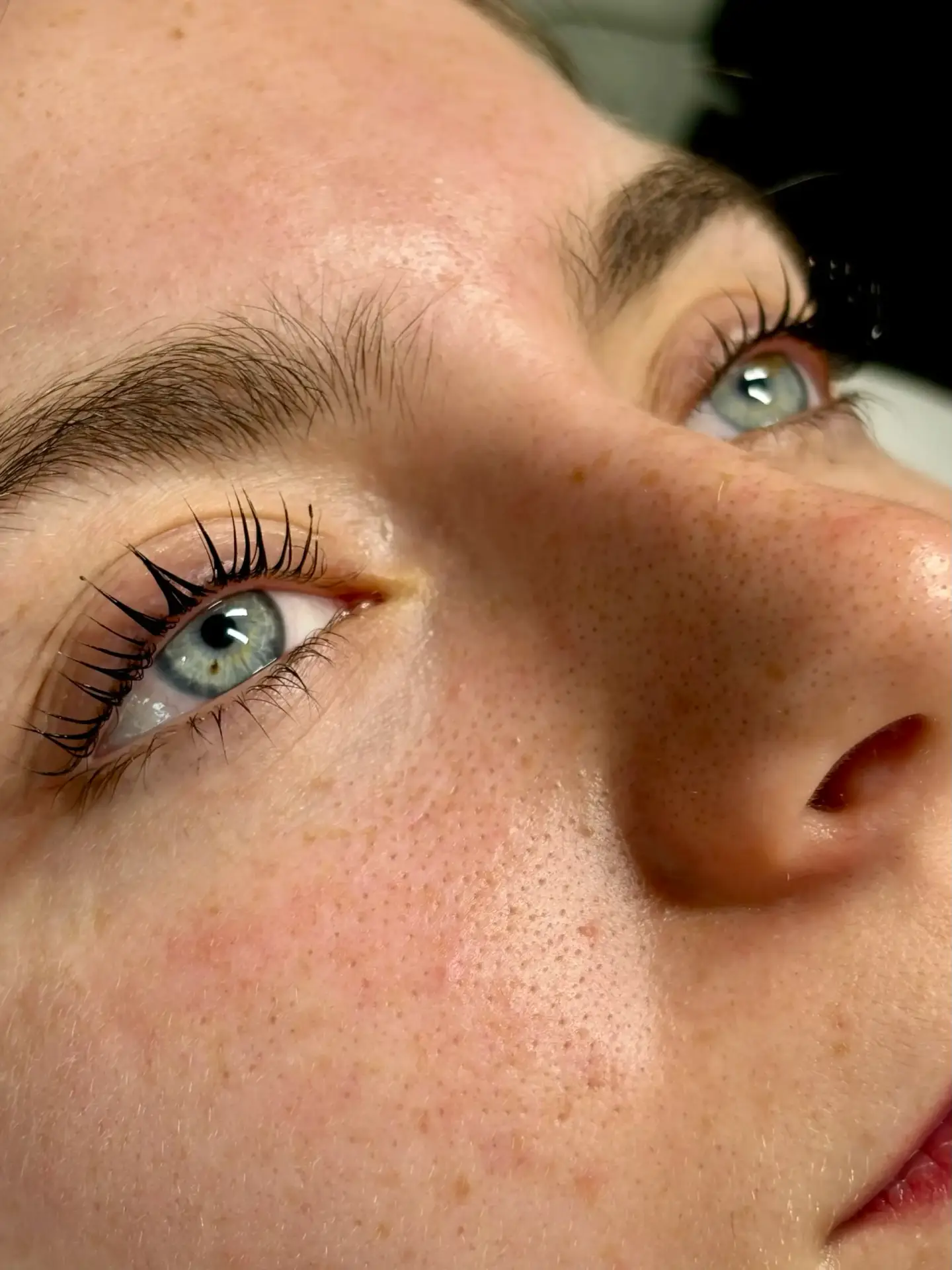 Close-up of a person's face showing blue eyes, long eyelashes, freckles, and smooth, light skin. The person is looking slightly upwards, and the image highlights facial details and texture.