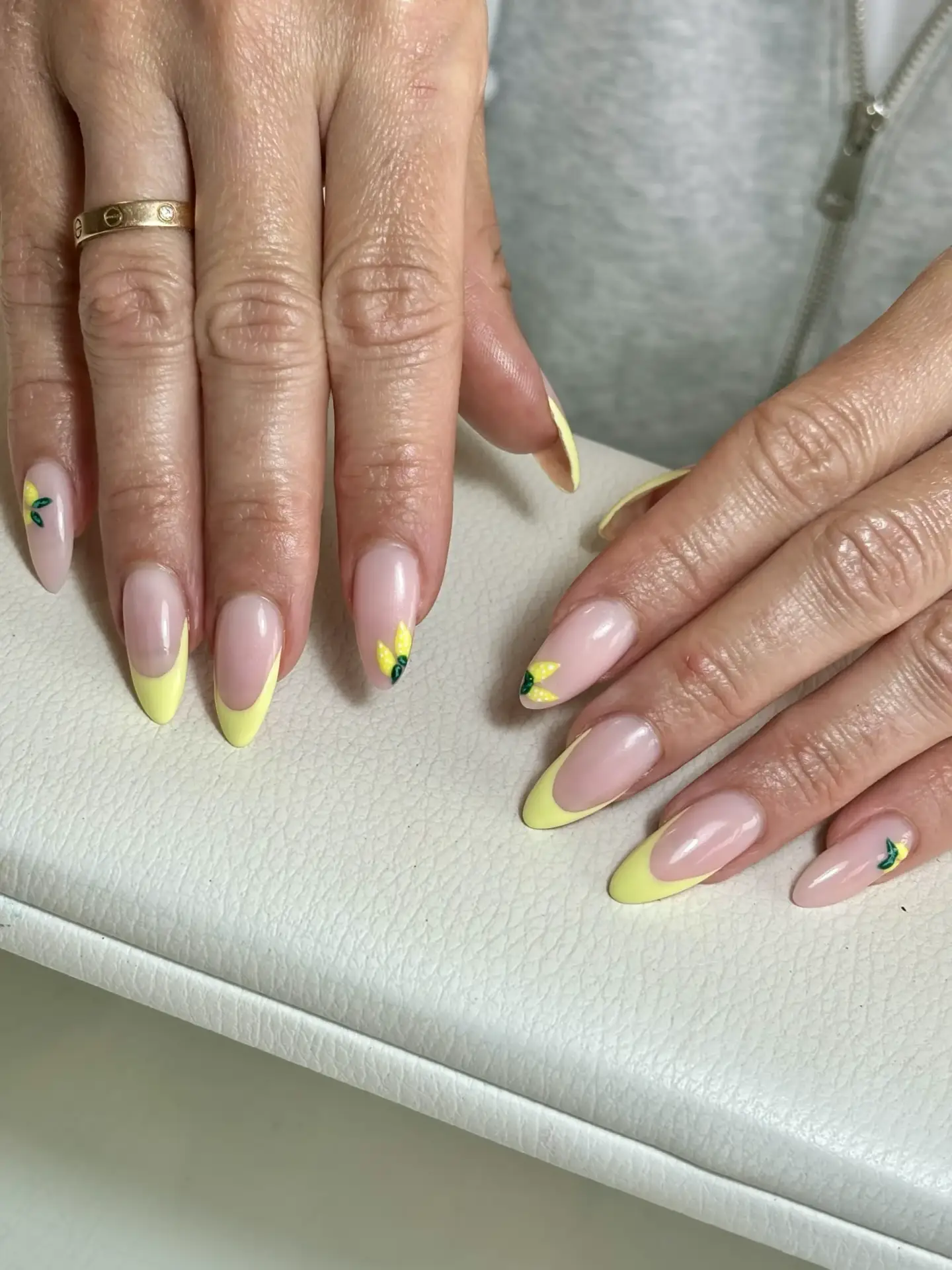 Hands with almond-shaped nails featuring a pale pink base, yellow French tips, and small lemon and leaf designs on selected nails, resting on a white textured surface.