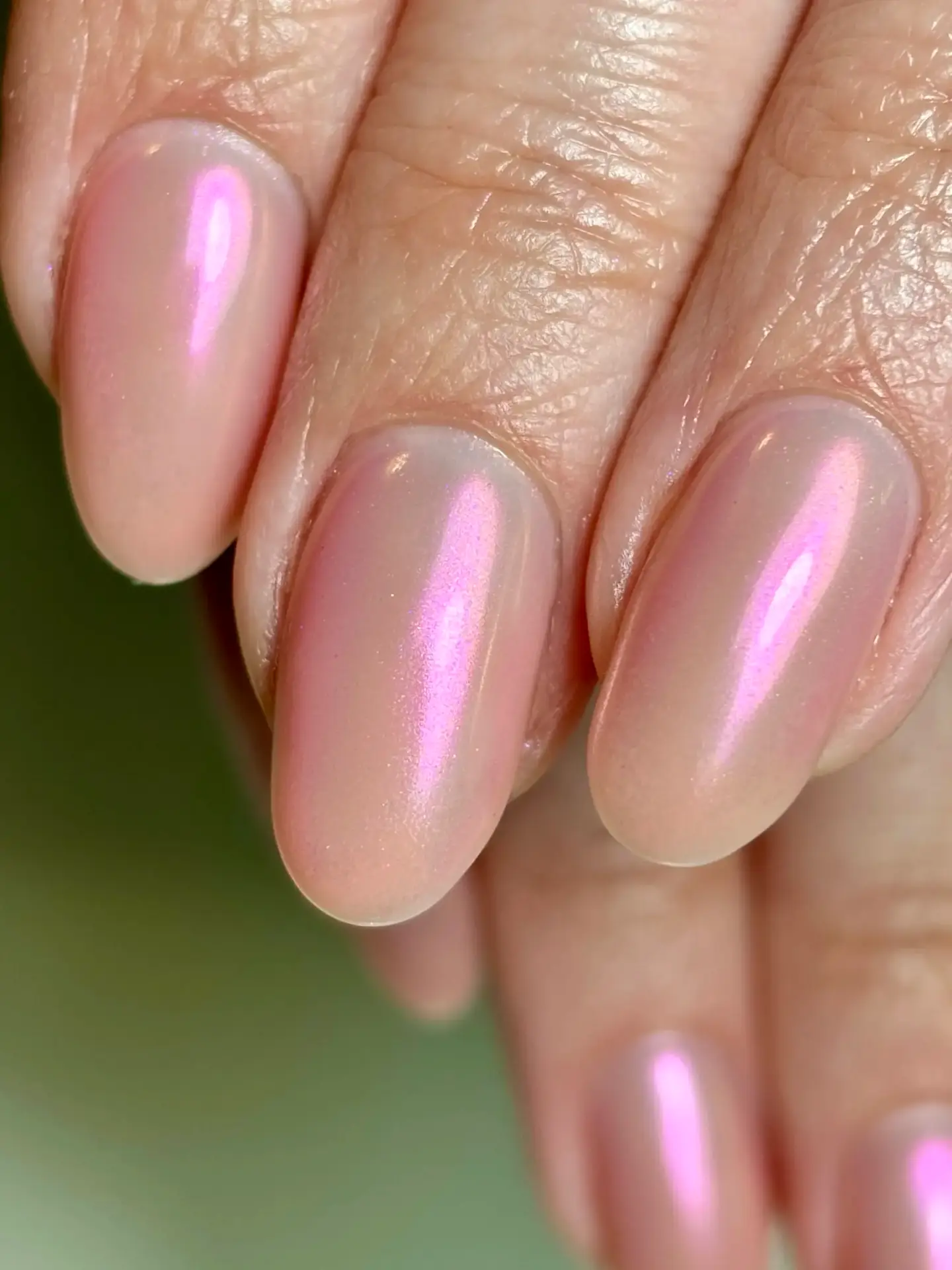 Close-up of a hand with almond-shaped nails painted in a shiny, iridescent light pink varnish. The nails have a smooth, glossy finish and the skin appears healthy.