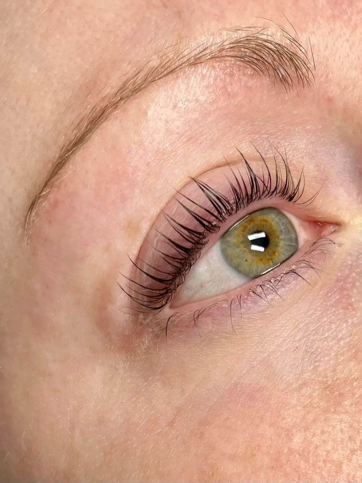 A close-up of a greenish-hazel eye with long, curled eyelashes and a well-groomed eyebrow on fair skin. The eye looks upward, showcasing details of the lashes and natural skin texture.