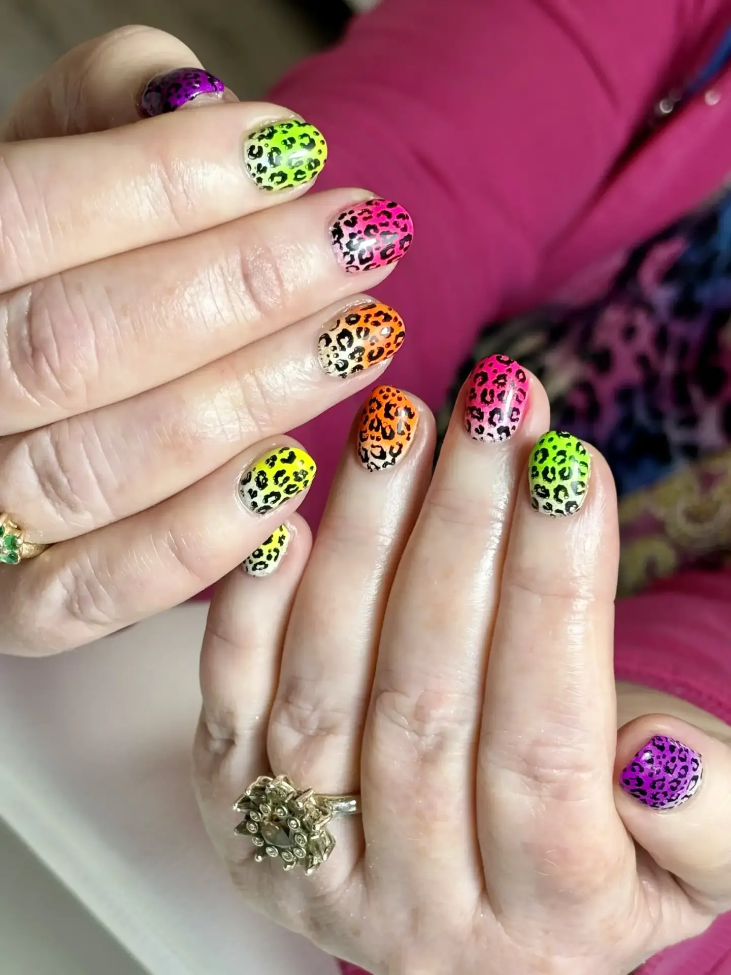 Hands with colourful leopard print nail art in neon shades of green, yellow, orange, pink, and purple. The person is wearing a gold floral ring and a pink jacket.