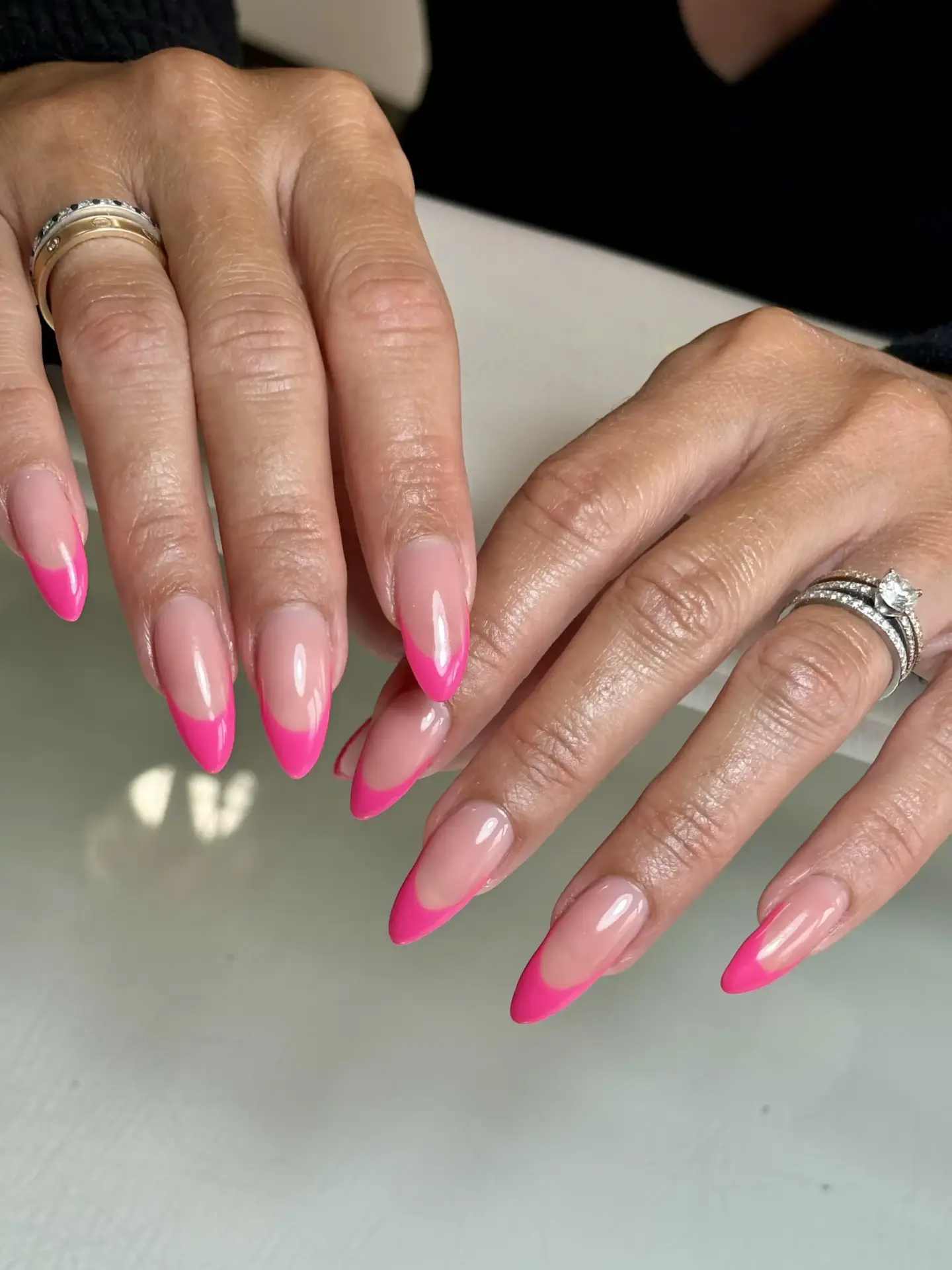A close-up of hands with long almond-shaped nails featuring a nude base and bright pink French tips. The person is wearing silver rings on several fingers.