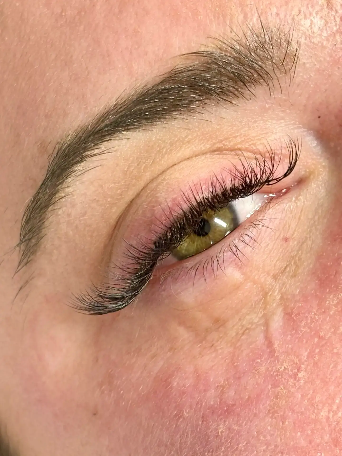 Close-up of a person’s eye with long, curled eyelash extensions, well-groomed eyebrow, and visible skin texture with some redness on the lower part of the face.