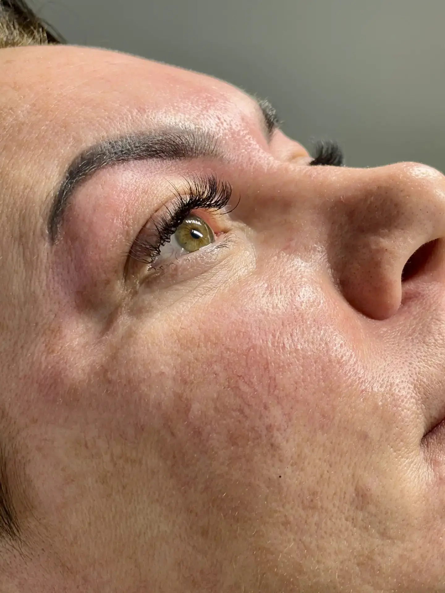 Close-up of a person’s face looking upward, showing detailed skin texture, light green eye, defined eyebrow, and long eyelashes. The image highlights the person’s natural features and complexion.