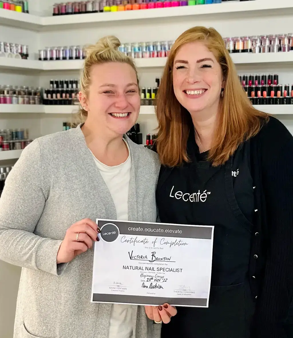 Two smiling women stand together in a nail salon, one holding a certificate of completion for Natural Nail Specialist. Shelves of nail varnish bottles are visible in the background.