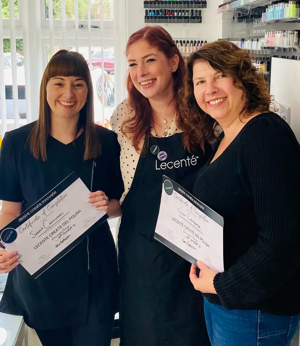 Three women stand indoors, smiling and holding certificates. Two women on either side wear black tops and hold certificates, while the woman in the middle, wearing an apron with Lecenté on it, stands between them. Shelves with nail varnish are visible behind them.