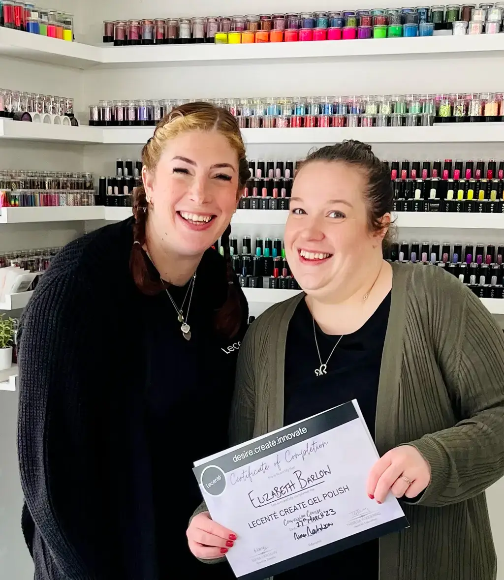 Two smiling women stand in front of shelves filled with colourful nail varnish bottles. One woman holds a certificate of completion for gel polish training, and both look happy and proud.