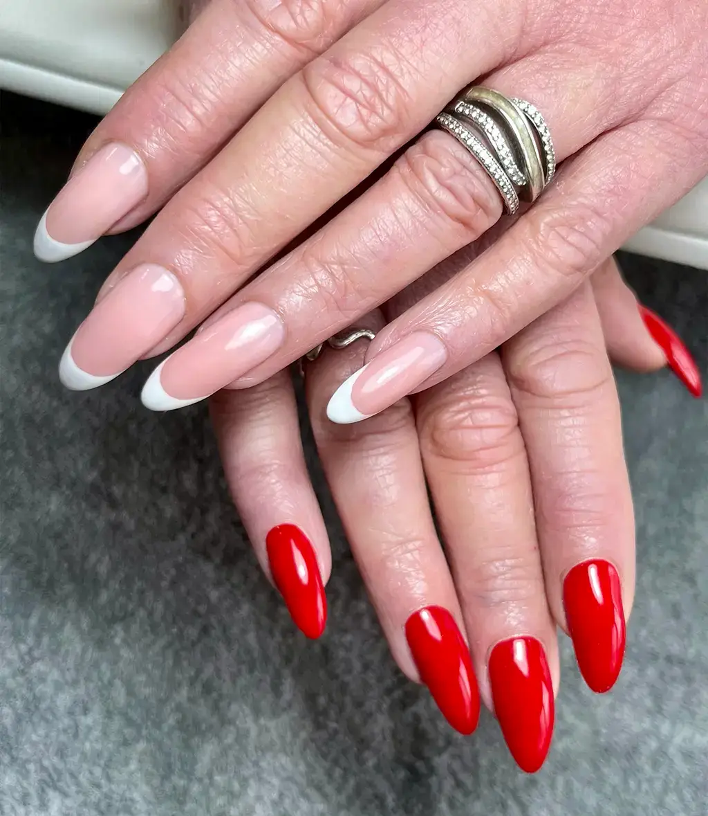 Two hands with manicured nails; one hand has classic French tips with nude and white varnish, while the other has glossy bright red nails. The person is wearing multiple silver rings.