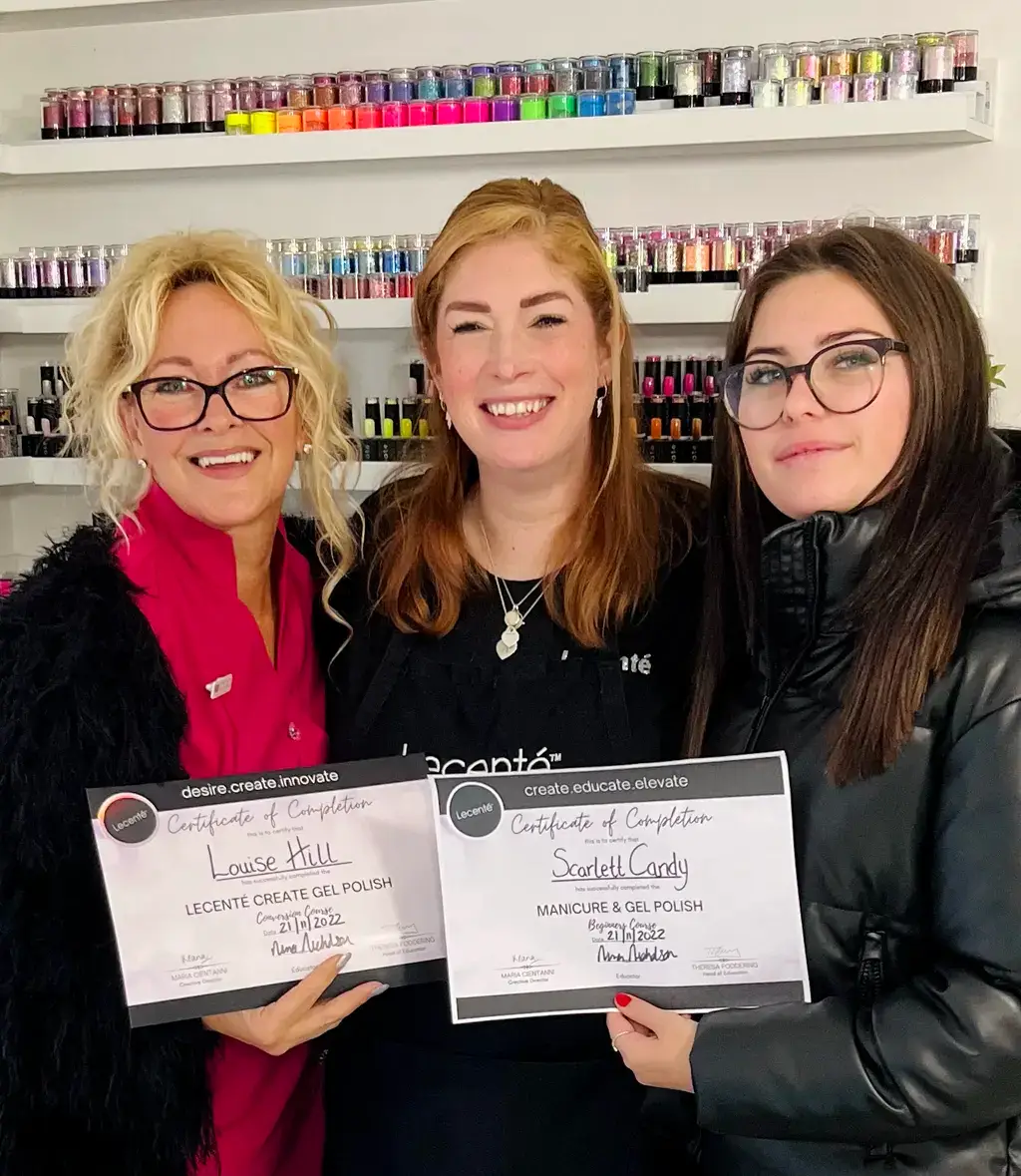 Three women stand smiling in a nail salon. Two women on either side hold certificates of completion for a gel polish course. Rows of nail varnish bottles are displayed on shelves in the background.