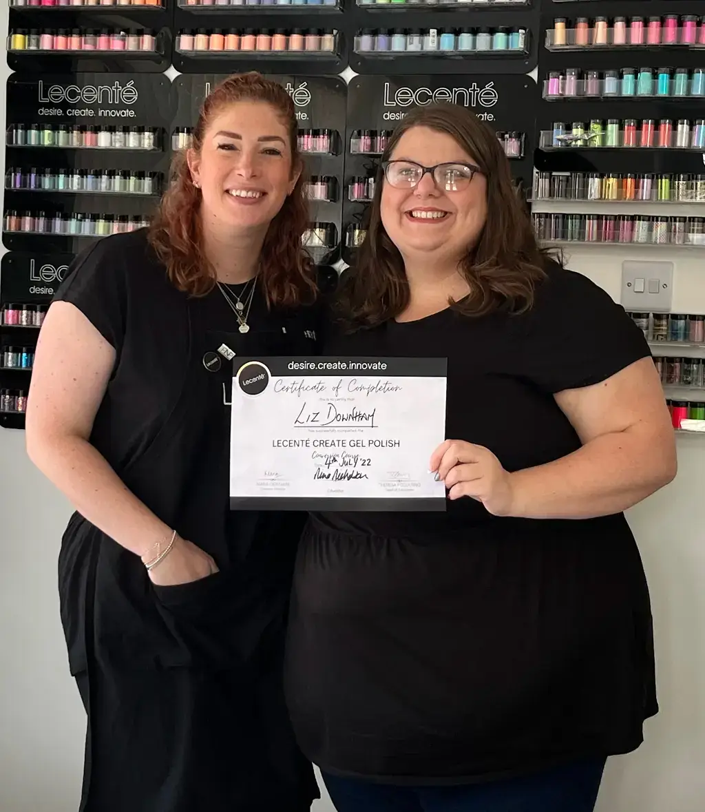 Two smiling women in black tops hold a Lecenté Certificate of Completion for Liz Downham’s Gel Polish course, standing before shelves of colourful nail products.