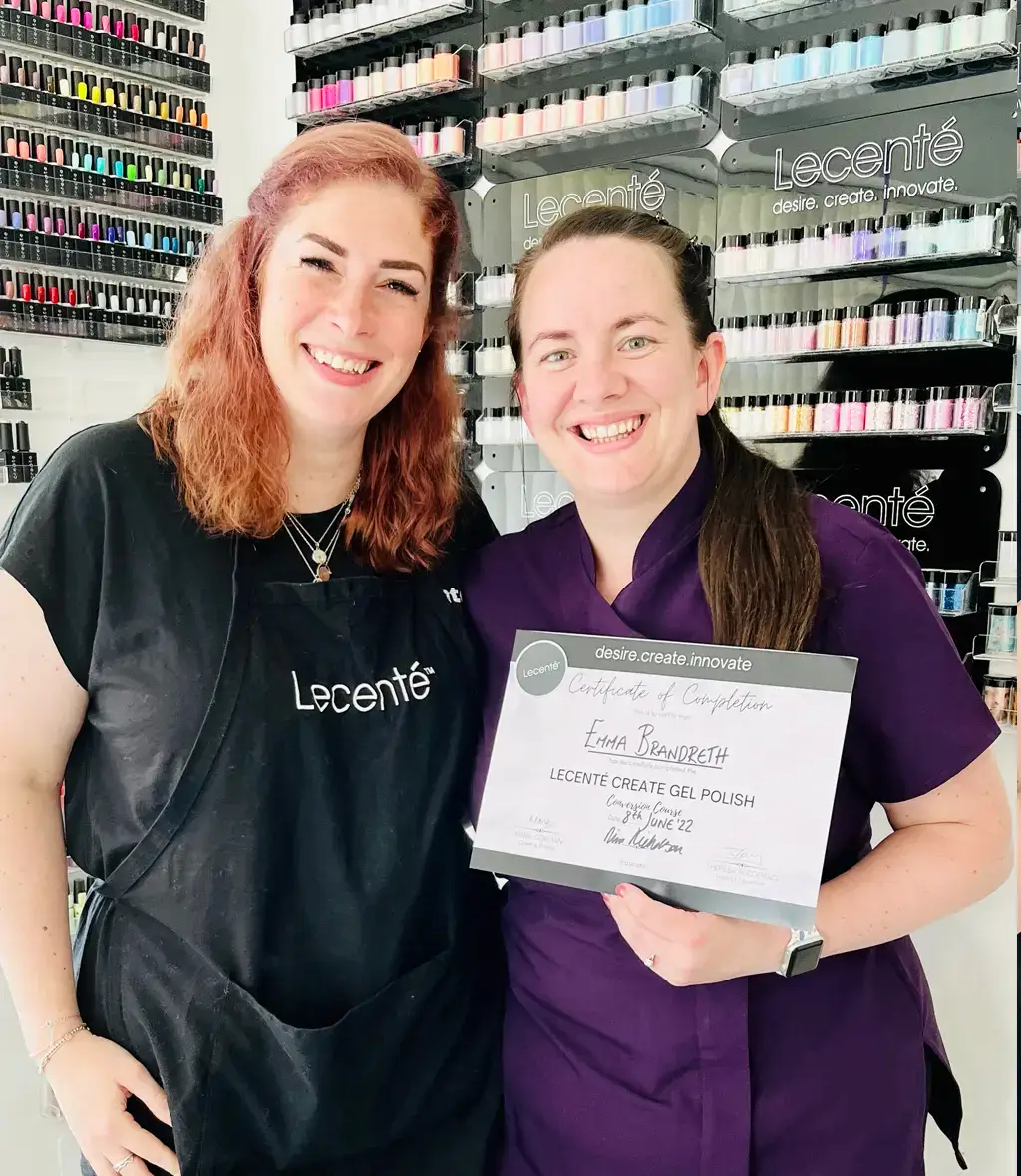 Two women stand smiling in a nail salon. One wears a black apron with Lecenté on it; the other holds a certificate of completion for a gel polish course. Colourful nail varnish bottles are displayed on the wall behind them.