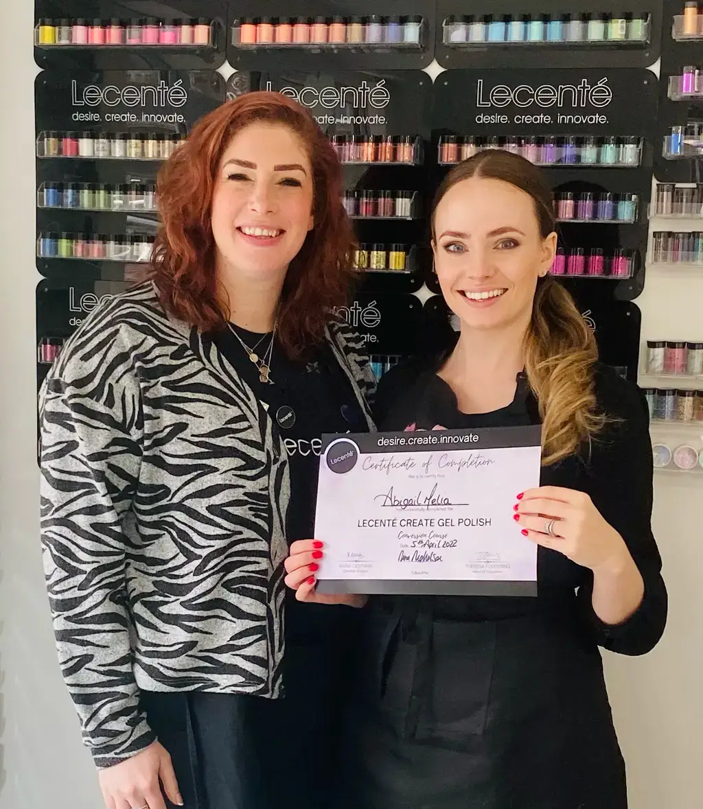 Two women stand smiling in front of a wall display of colourful nail powders. One woman holds a certificate for completing the Lecenté Create Gel Polish course.