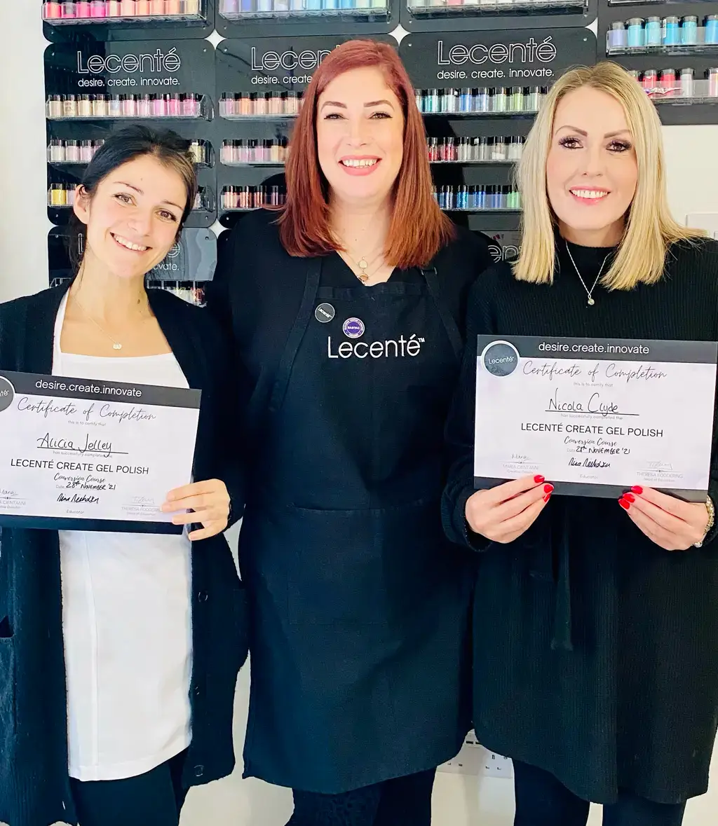 Three women stand smiling indoors. The woman in the centre wears a black apron with the Lecenté logo. The two women on either side hold certificates of completion for Lecenté Create Gel Polish. Shelves of nail products are in the background.