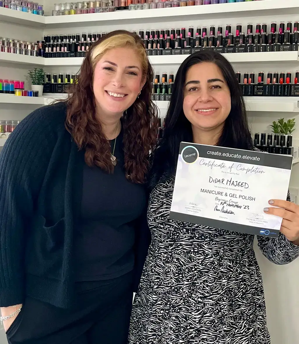 Two women stand smiling in a nail salon. One woman holds a certificate of completion for manicure and gel polish. Shelves with colourful nail varnish bottles line the wall behind them.