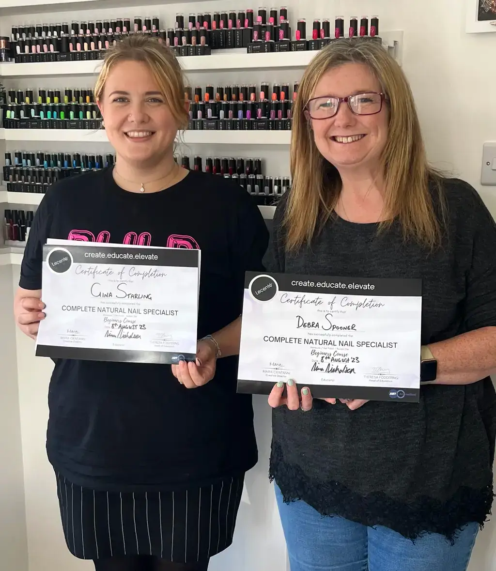 Two smiling women stand side by side holding certificates in front of a wall of nail varnish bottles. Both certificates are titled “Complete Natural Nail Specialist.”.