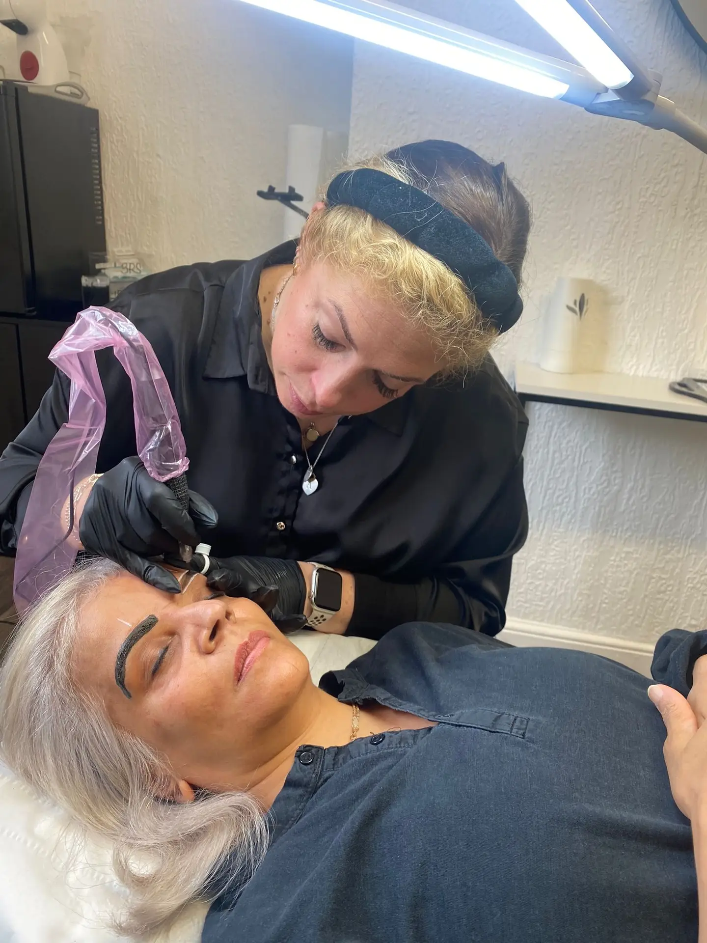 A beauty therapist wearing gloves and a black headband carefully applies permanent make-up to the eyebrows of a woman lying down with her eyes closed in a salon setting.
