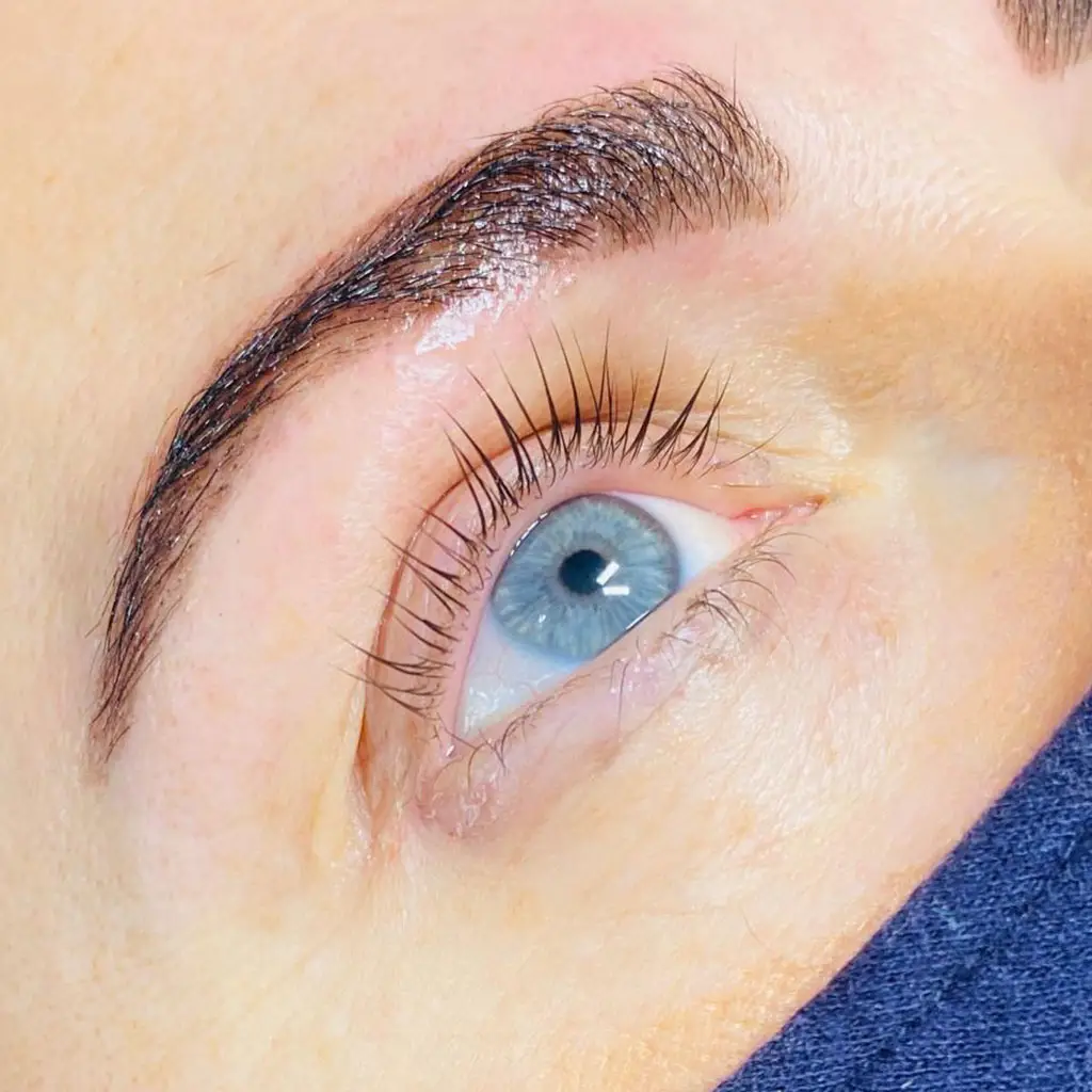 Close-up of a blue eye with long, curled eyelashes and a well-groomed eyebrow. The skin is light, and part of a dark face mask is visible at the bottom right corner.