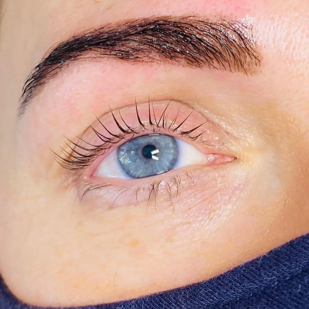A close-up of a blue eye with long, curled eyelashes and a well-groomed eyebrow. The skin appears fair, and part of a dark face mask is visible at the bottom of the image.