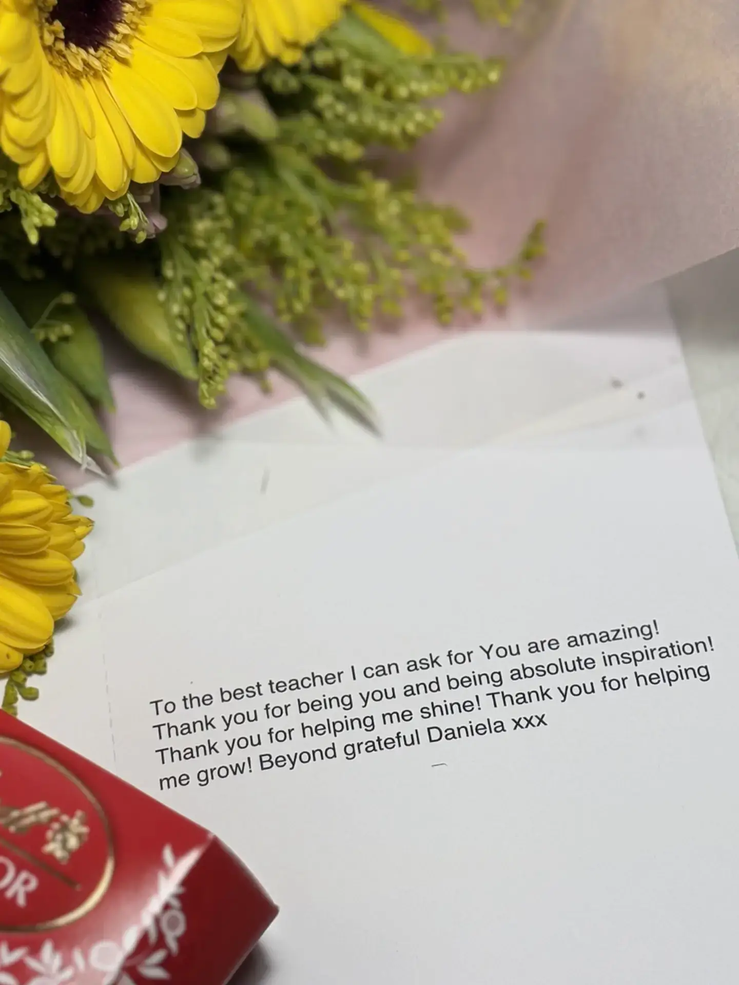 A bouquet of yellow flowers and a card with a heartfelt thank you message for a teacher, resting on a white surface next to a red Lindor chocolate box.